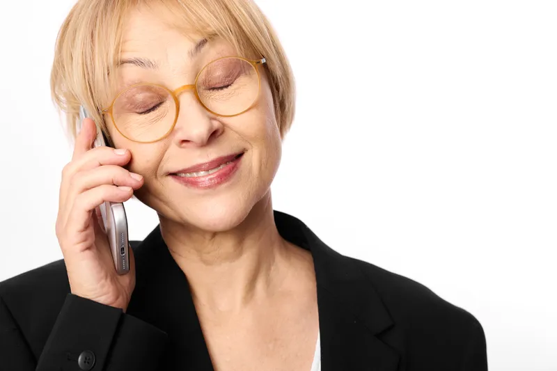 Woman with glasses smiling while speaking on smartphone for phone call, communication and business portrait, studio light, clean white background, mature blonde hair