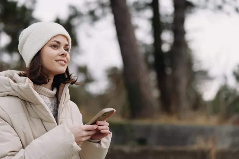 Woman in a winter outdoor setting uses a phone, wearing a jacket and beanie, a cozy scene with trees and a calm park atmosphere, natural light and focused mood in a chilly day