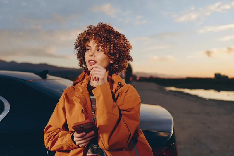 Woman lifestyle portrait near a car at sunset, curly hair, orange jacket, holding a phone, relaxed smile, outdoors on a road, soft evening light, travel mood