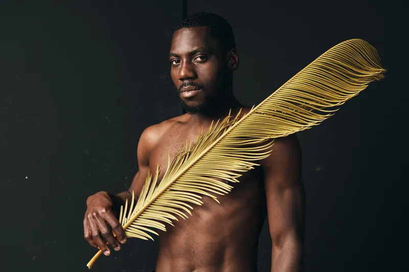 Black man with dark skin holding large golden palm leaf in creative visual designer style, artistic portrait with bare torso and serious expression on black background.