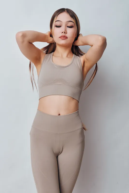 Young woman in beige sportswear stretching with hands behind head, eyes closed, standing against neutral background for fitness and wellness. Relaxed, healthy lifestyle and calm pose.