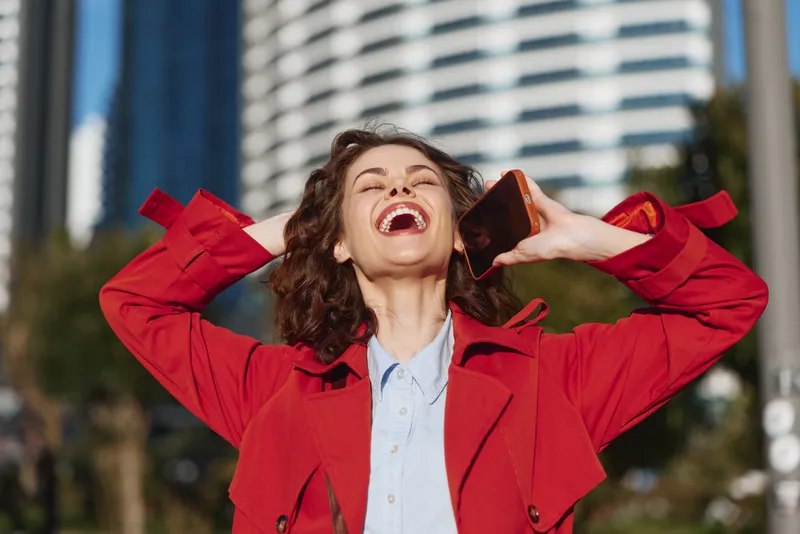 A joyful woman in a red coat laughs while talking on a smartphone in an urban outdoor setting, with a modern city backdrop, conveying energy and spontaneous connection.