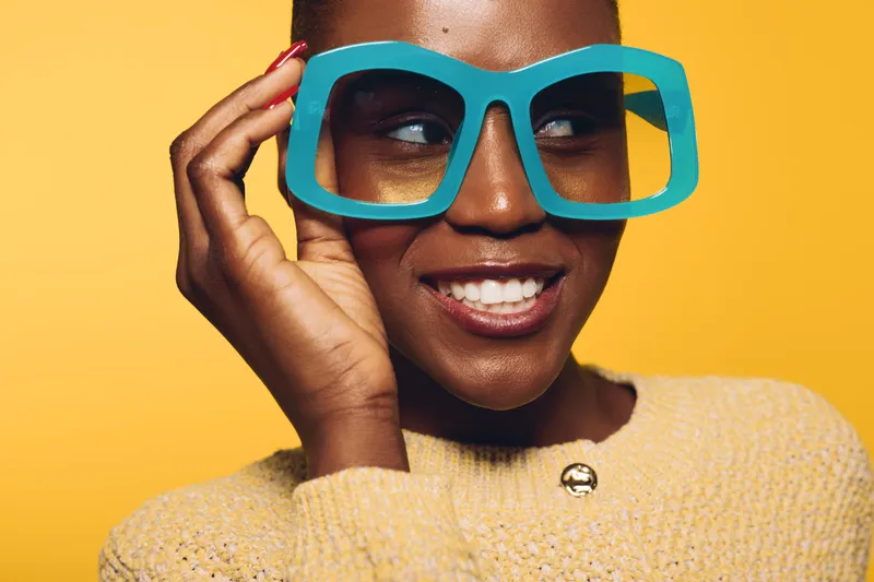 Smiling black woman with dark skin wearing oversized blue glasses and yellow sweater looks aside on yellow background. Concept for fashion, style, and joyful mood with vibrant colors.