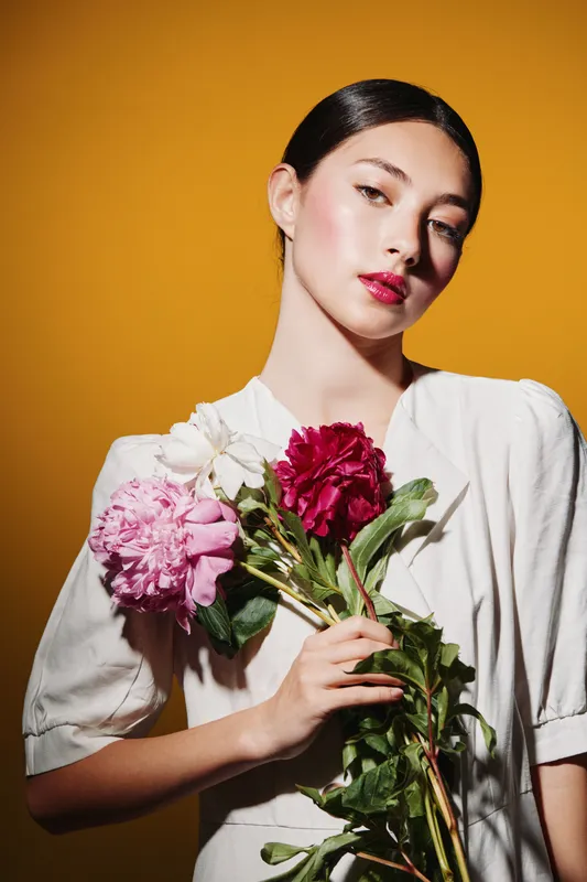 Young fashion model with glossy red lips and sleek hair holds vibrant peonies against orange background. Creative beauty portrait with soft natural makeup and elegant styling.