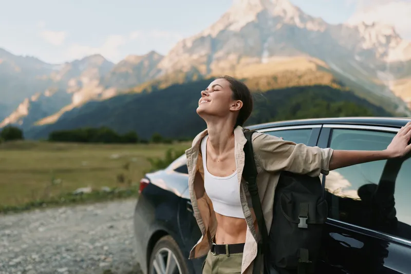 Young woman in casual outfit leans on a car door with eyes closed enjoying fresh mountain air during a sunny day in nature, expressing relaxation and freedom.