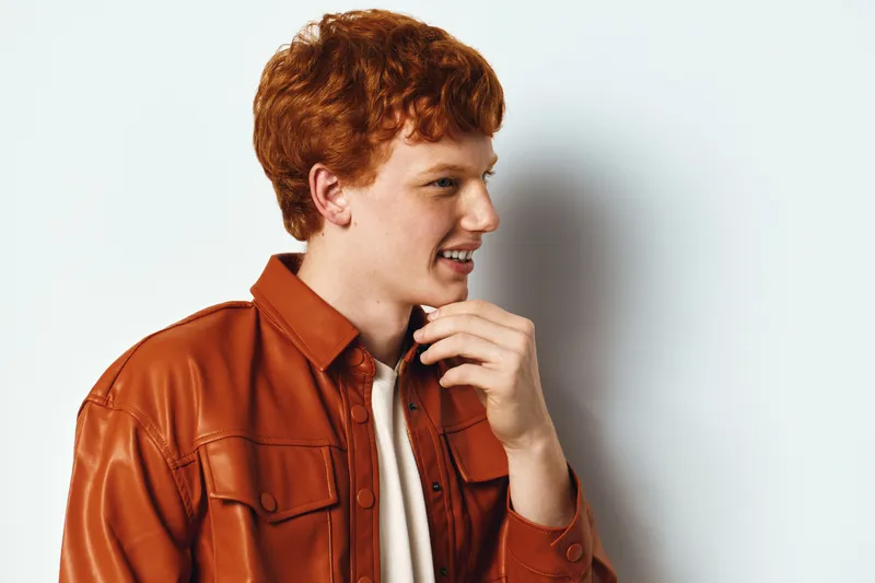 Redhead man with curly hair wearing brown jacket standing and turning sideways looking away with thoughtful expression against white background. Casual style portrait.