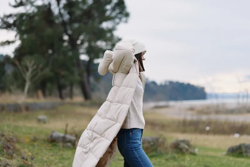 Walking outdoors, a person in a long coat and puffer jacket walks through a wide landscape with a field and distant water, conveying a calm winter lifestyle and natural scene.