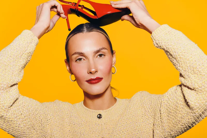 Young woman with slicked hair holds red high heel shoes above her head against bright yellow background. Fashion concept with confident model wearing beige sweater and gold hoop earrings.