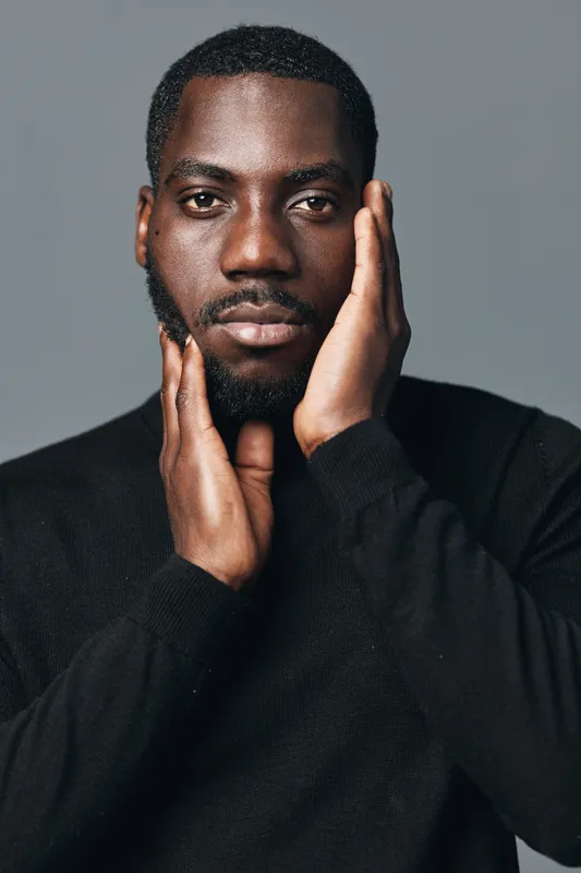 African man with dark skin and beard posing with hands on face in creative conceptual visual style. Portrait of confident male model wearing black sweater on gray background.