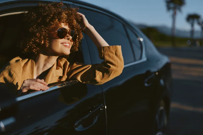 Smiling woman with curly hair leaning out of a car window, wearing a jacket and sunglasses. Retro film color palette enhances warm vintage mood and carefree travel vibe on the road.
