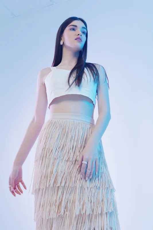 Young woman in a white crop top and fringed skirt poses confidently in soft studio lighting. Concept for beauty, fashion, and modern style with elegant minimalism.