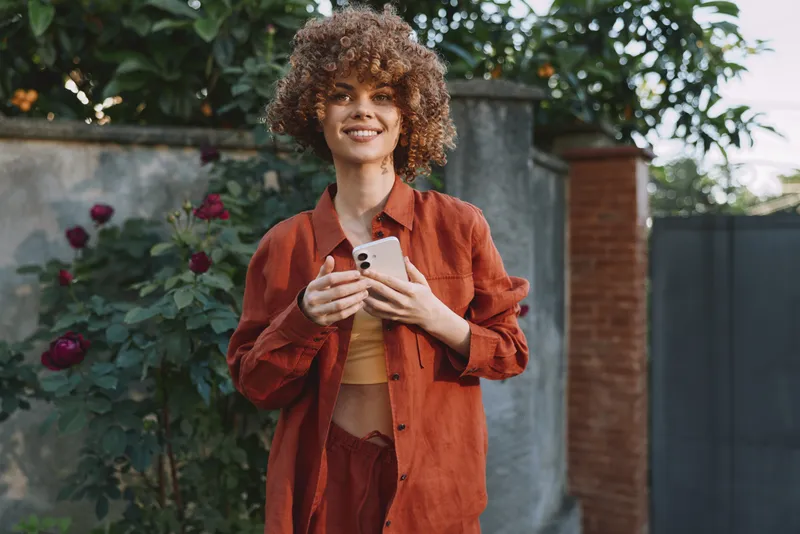 Smiling woman with curly hair holding a smartphone outdoors in a rust orange outfit. Close, casual portrait highlighting fashion, lifestyle, and self expression