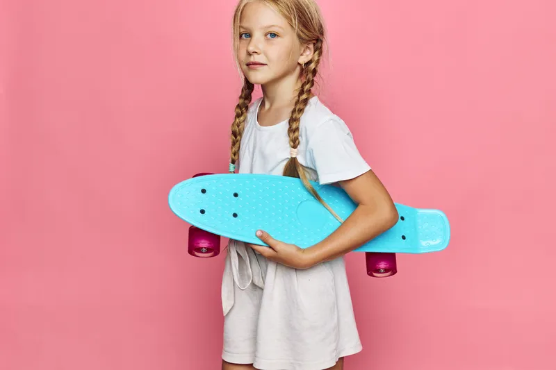 Young girl with braids holds a blue skateboard while standing against a pink background. She wears a white t-shirt and beige shorts. Concept of childhood and outdoor fun.