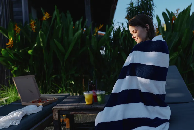 Relaxed woman outdoors, wrapped in a striped blanket, enjoying a sunlit moment, casual lifestyle, garden setting, refreshing drinks nearby, snack on the table, natural colors, peaceful mood