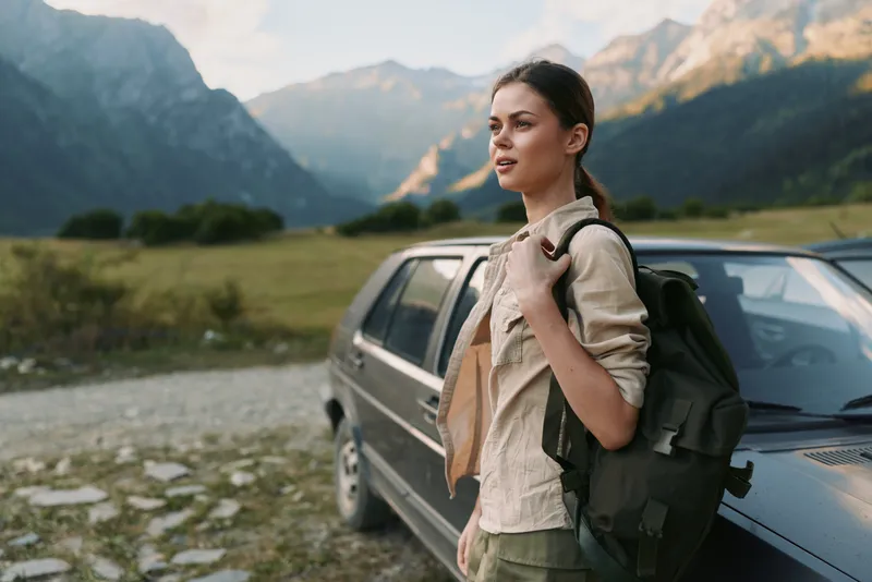 Smiling woman with backpack beside a car in a valley with mountains in the background, wearing a casual jacket, enjoying an outdoors lifestyle, travel and adventure vibe under natural light.