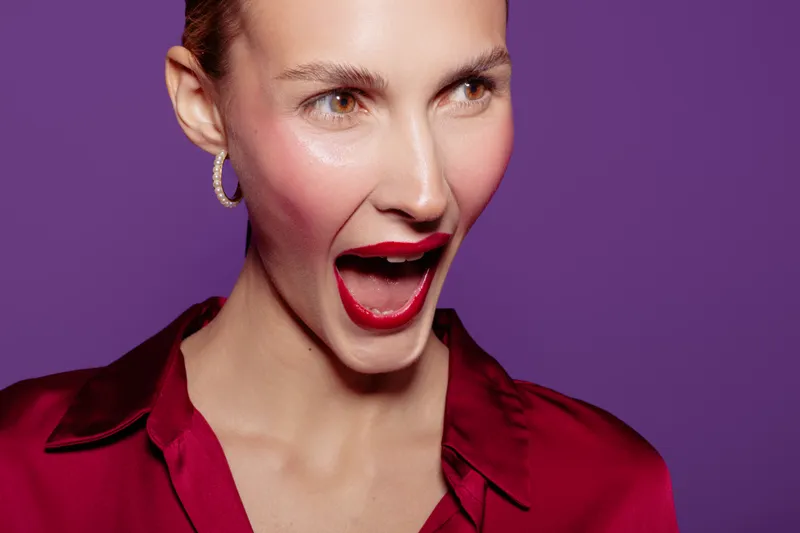 Close-up of excited woman with red lipstick and earrings wearing red blouse on purple background. Concept of beauty, fashion, makeup, and expressive emotions in studio lighting.