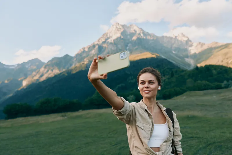 Young woman lifestyle taking selfie with smartphone outdoors, mountain landscape background, casual outfit, natural light, smiling face, summer day, travel concept