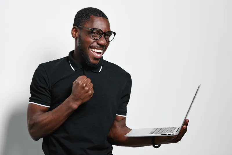 African American man with dark skin and glasses holding laptop and celebrating success with fist pump in casual black shirt on white background. Happy, excited, technology user.