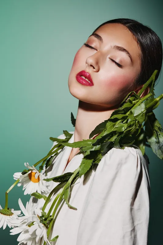 Close-up of serene woman with perfect makeup, glossy lips and natural floral accessories. Fashion beauty concept highlighting creative style with fresh blooming daisies on green background.