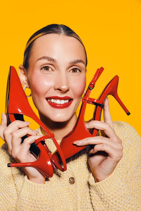 Smiling woman with red lipstick holds red high heel shoes near face on bright yellow background. Concept of fashion, style, footwear, and joyful emotions in studio portrait.