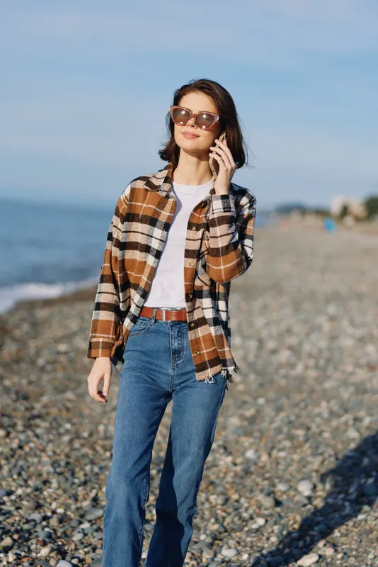 Casual woman on a pebble beach talking on a mobile phone, wearing a brown plaid shirt, white tee and blue jeans, sunglasses, relaxed pose with sea in the background.
