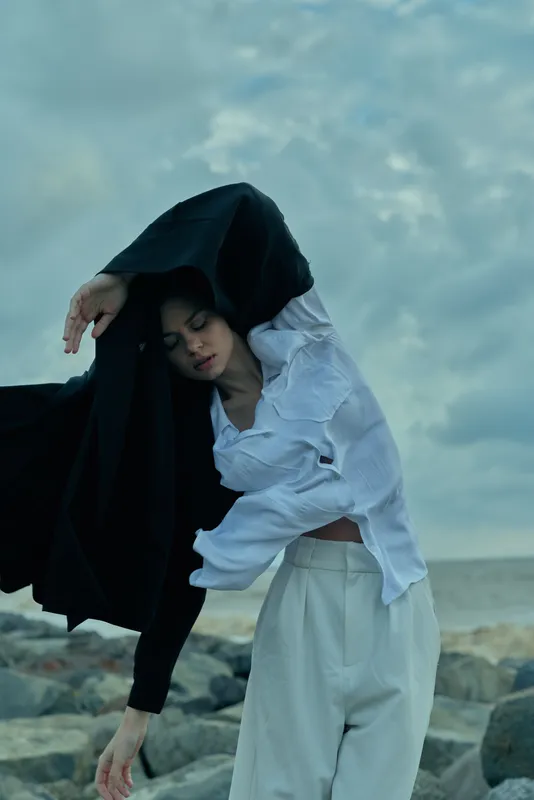 Young adult woman on a windy rocky coastal shore wearing a loose white shirt and a dramatic black scarf flowing with the sea breeze in a fashion editorial mood atmosphere