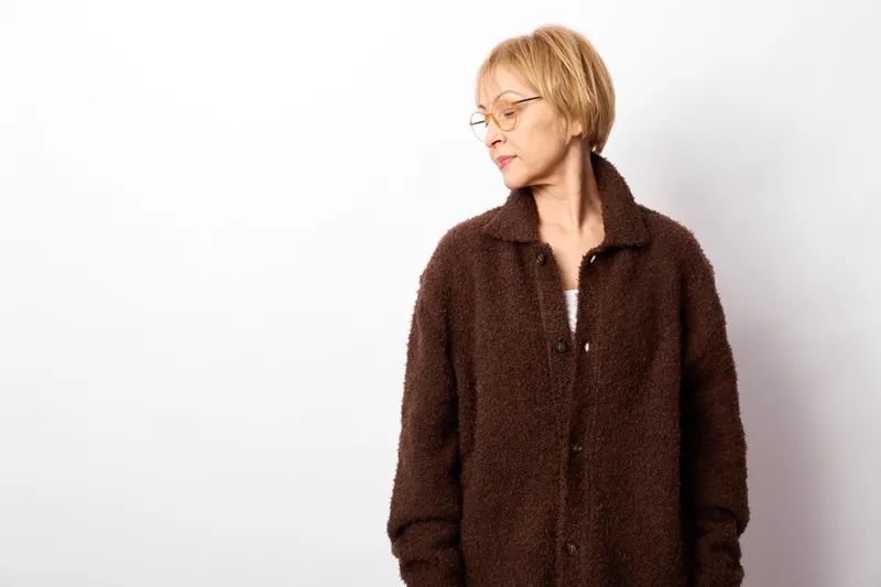 Woman portrait, mature adult wearing brown coat, short blond hair, eyeglasses, standing in studio against white background, calm expression, fashion lifestyle, side view