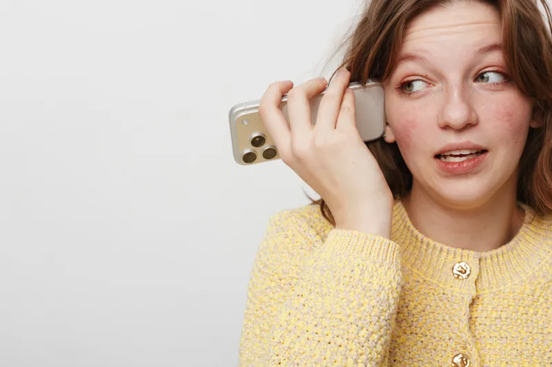 Young woman in yellow sweater holds smartphone to ear, looking aside with uncertain expression. Concept of communication, hesitation, and casual lifestyle in modern technology use.