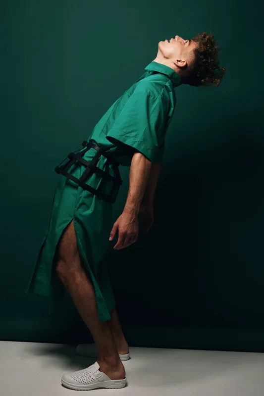 Young man in green outfit with side slit and black belt posing with head tilted back against dark background. Fashion model in casual shoes showing dynamic posture and curly hair.