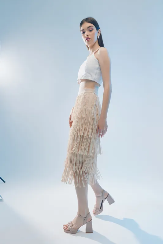 Slim woman beauty fashion model poses in beige fringe skirt and white top on light background. Studio shot with soft lighting. Concept for style, elegance, and modern trends.