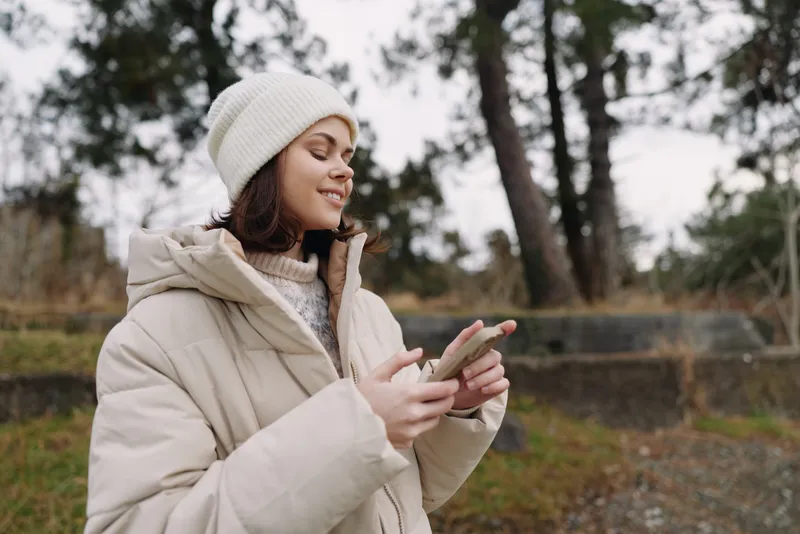 Woman in a warm beige coat and knit hat using a smartphone outdoors among trees, capturing a candid winter moment with quiet focus, soft light and a calm natural backdrop