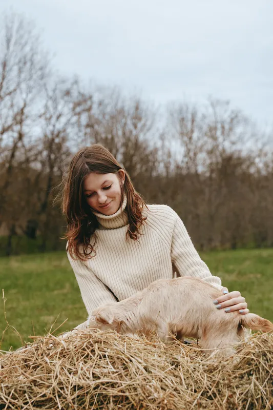 Young woman in a beige sweater gently pets a light brown baby goat resting on a haystack in a green field during an overcast day in early spring.