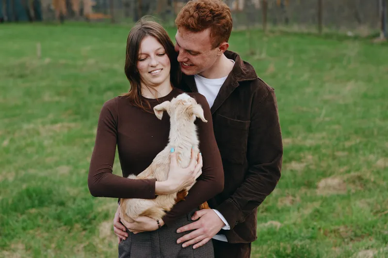 Couple outdoors lifestyle portrait, goat kid held by a smiling pair in a cozy scene, nature park setting, candid affection, warm light, gentle connection, rustic attire, joyful moment in a serene