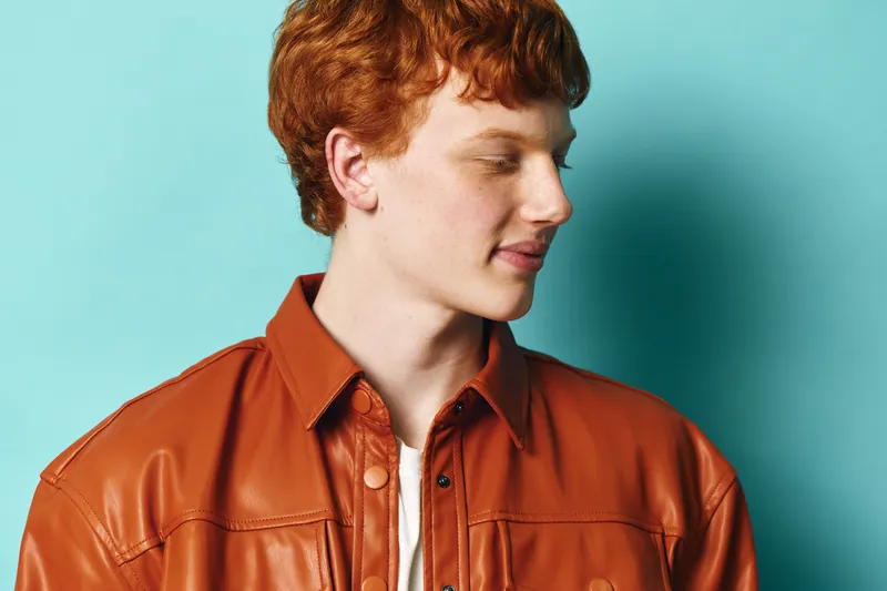Young man with red hair and freckles wearing a brown leather jacket looks sideways against a blue background in a studio portrait, casual style and thoughtful expression.