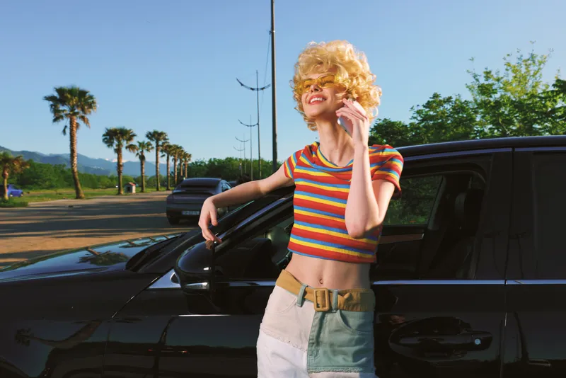 A young woman with curly blonde hair in a striped crop top leans against a dark car in a sunny lot, exuding casual confidence and a retro film color vibe.