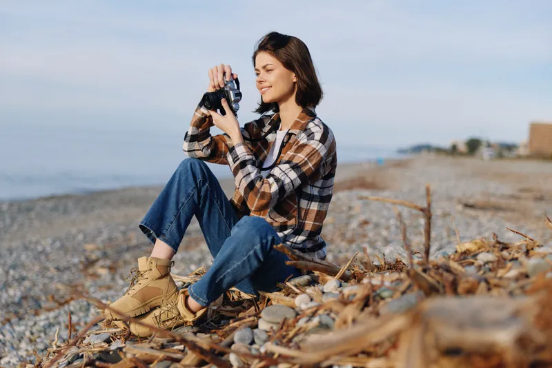 Woman sitting on a rocky beach with a camera, capturing a moment outdoors. Casual plaid shirt and boots among autumn leaves for a calm, coastal scene with natural light