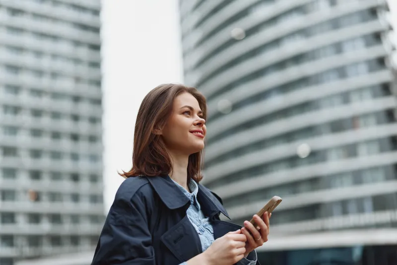 Young woman with brunette hair smiles while holding a smartphone outdoors near a modern curved glass building on a cloudy day.