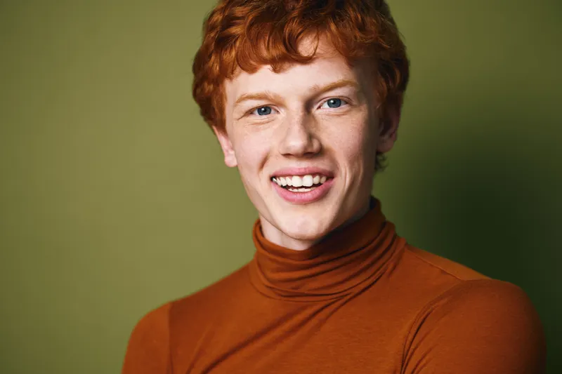 Young man with red hair and freckles smiling wearing brown turtleneck sweater on green background. Portrait of confident caucasian male with positive expression.
