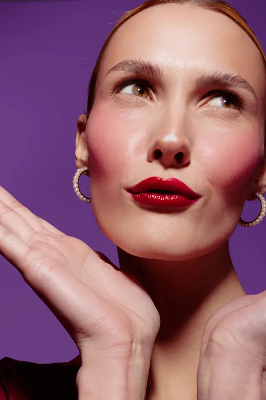 Close-up of a woman with red lipstick and hoop earrings posing with hands near face on purple background. Concept of beauty, makeup, fashion, and confident expression.