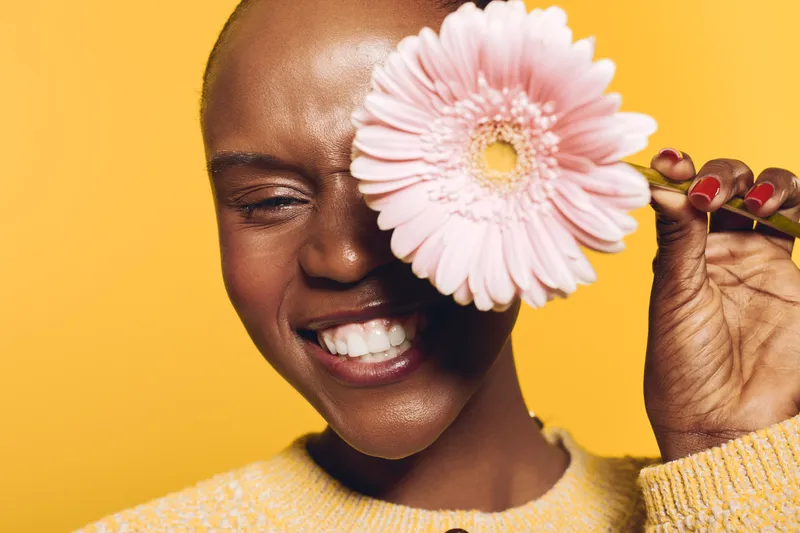 Smiling dark skinned African woman with short hair holds pink flower near face against yellow background. Concept of natural beauty, joy, floral, and positive emotions in studio portrait.
