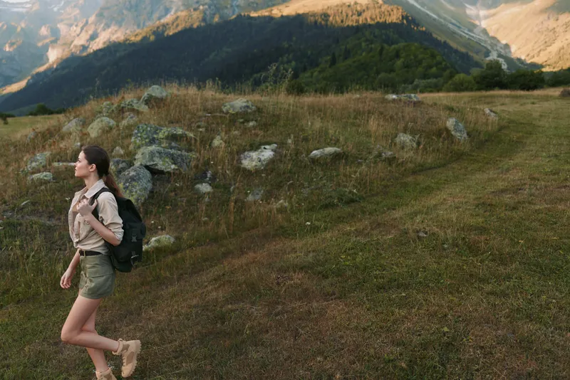 Cheerful woman hiking outdoors with backpack, casual attire, grassy trail, mountain ridge in background, natural light, energetic lifestyle, scenic nature moment outdoors