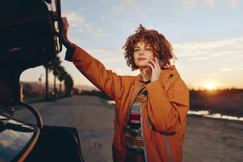 Woman outdoors, car, jacket, sunset, lifestyle, curly, hair, cheerful expression, standing by vehicle, adjusting coat collar, warm golden hour light, open road background, travel mood