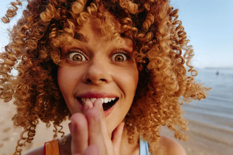 Close-up of a young woman with curly hair on a sunny beach, playfully sticking out her tongue at the camera with the sea in the background, her bright expression and the warm sunlight creating a