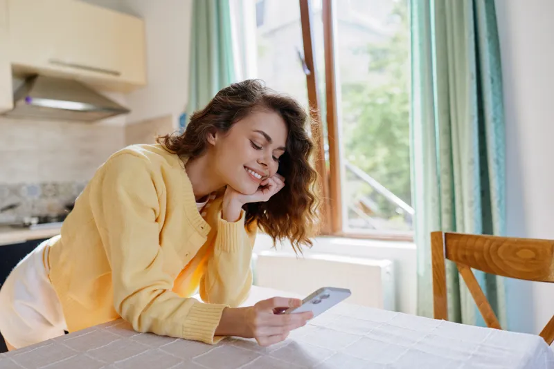 A young woman in a yellow sweater relaxes at a bright kitchen table, smiling softly as she looks at her phone, creating a cozy, home moment.