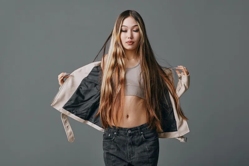Young woman with long hair wearing casual jacket and crop top posing with open arms on gray background. Fashion, style, confidence, beauty, and trendy outfit concept.
