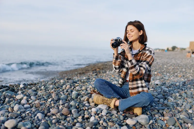 Woman photographer sits on a pebble beach holding a camera and smiling as waves roll in Casual outdoor scene capturing nature travel vibes seaside memories and creative storytelling