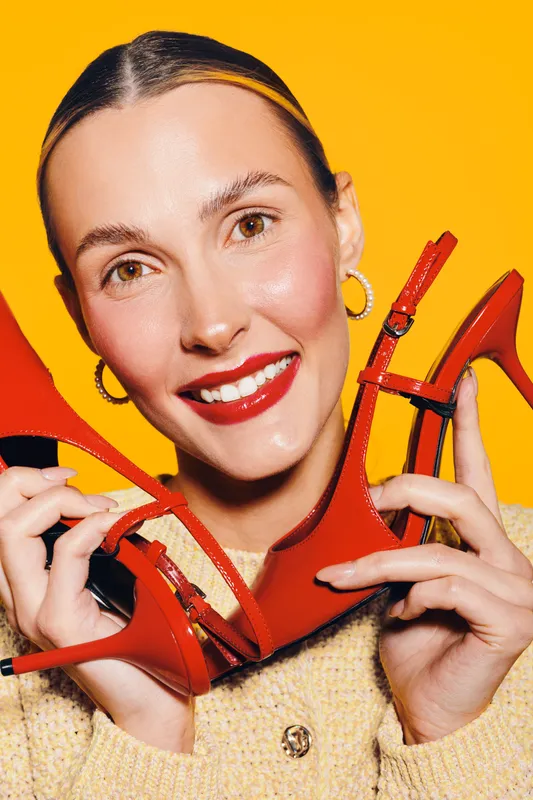 Smiling woman with red lipstick holds red high heels near her face on bright yellow background. Concept of fashion, beauty, style, and vibrant accessories for modern lifestyle.