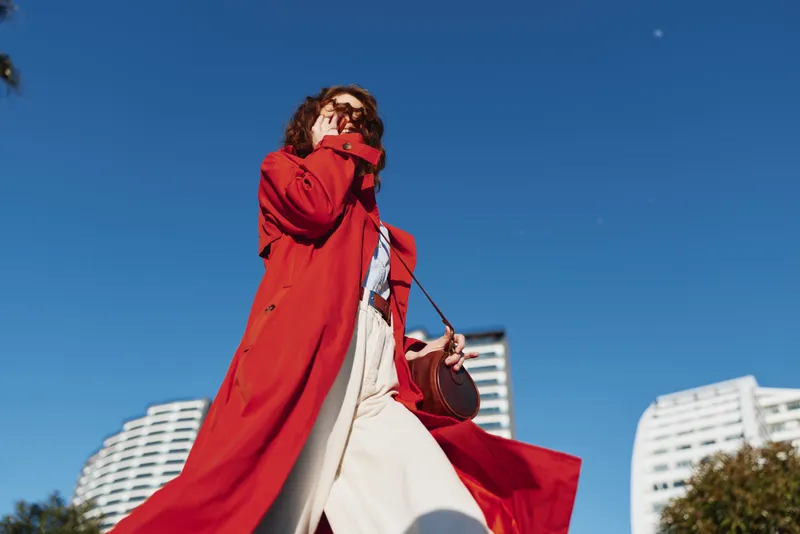 A single brunette woman in a bright red coat strides forward in a clear blue sky, urban buildings visible behind her, conveying movement and confidence.