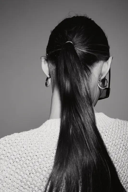Portrait of an Asian woman with long black hair tied in a ponytail wearing hoop earrings and a knitted sweater seen from behind in black and white studio setting.