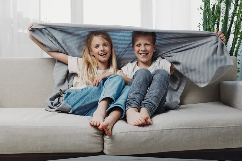 Happy children sitting barefoot on sofa covered with blanket, smiling and enjoying cozy home moment in bright living room with natural light and plants.